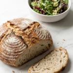 Sauerteig-Bauernbrot serviert mit bunter Blattsalat — Eine abgeschnittene, saftige Brotscheibe liegt neben knackigem Salat.