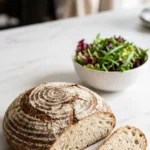 Roggenmischbrot mit Sauerteig serviert mit bunter Blattsalat — Eine abgeschnittene, saftige Brotscheibe mit krosser Kruste liegt einladend neben frischem Salat.