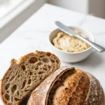 Dinkel-Sauerteigbrot serviert mit Kräuterbutter — angeschnittener Laib zeigt die saftige, offen porige Krume.