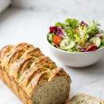 Kräuterbaguette serviert mit bunter Blattsalat — Ein abgebrochenes Stück des saftigen Ährenbrotes liegt appetitlich neben frischem Salat.