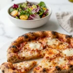 Pizza aus Sauerteig serviert mit bunter Blattsalat — Ein abgeschnittenes Stück Pizza liegt einladend neben einer Schale Salat.