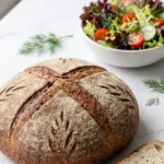 Polnisches Roggenbrot serviert mit bunter Blattsalat — Eine abgeschnittene, saftige Brotscheibe mit Kümmelkörnern neben frischem Salat.