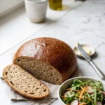 Roggenbrot ohne Sauerteig serviert mit bunter Blattsalat — Eine abgeschnittene, saftige Brotscheibe mit krosser Kruste liegt neben knackigem Salat.