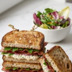 Sauerteig-Sandwich serviert mit bunter Blattsalat — Ein halbiertes Speck-Sandwich liegt appetitlich neben einer Schale Salat.