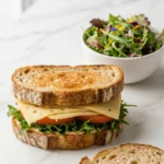 Sauerteigbrot-Sandwich serviert mit bunter Blattsalat — Ein halbiertes Käsesandwich liegt neben einer Schale frischem Salat.