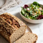 Flockenbrot mit Sauerteig serviert mit bunter Blattsalat — Eine abgeschnittene, saftige Vollkornbrotscheibe liegt appetitlich neben frischem Salat.
