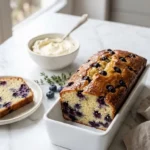 Zitronen-Blaubeer-Brot serviert mit Mascarpone-Creme — saftig angeschnittene Scheibe auf elegantem weißem Marmor.