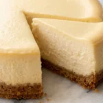 New York Cheesecake frisch gebacken — feine goldgelbe Oberfläche und knuspriger Keksboden auf weißem Marmor.