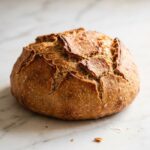 Saftiges Sauerteig-Roggenmischbrot frisch gebacken — rundes, krustiges Landbrot mit natürlichem Ausbund auf weißem Marmor.