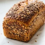 Leinsamenbrot frisch gebacken — kerniges, dunkles Kastenbrot überstreut mit reichlich Saaten auf weißem Marmor.