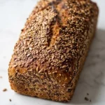 Dinkel-Buchweizen-Brot frisch gebacken — kerniges dunkles Kastenbrot überstreut mit reichlich Saaten auf weißem Marmor.