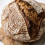 Bauernbrot frisch gebacken — runder, rustikaler Brotlaib mit tiefer Kruste und Mehlmuster auf einem Holzbrett vor Marmor