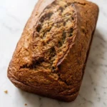 Sauerteig-Bananenbrot frisch gebacken — saftiges, dunkel-goldbraunes Kastenbrot mit feiner Kruste auf weißem Marmor.