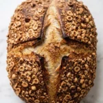Haselnussbrot frisch gebacken — rundes rustikales Sauerteigbrot überstreut mit gerösteten Haselnüssen auf weißem Marmor.