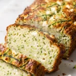 Herzhafter Zucchinikuchen frisch gebacken — pikantes dunkel-goldbraunes Kastenbrot mit Feta und Parmesan auf weißem Marmor.