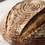 Roggenbrot frisch gebacken — kräftiges Sauerteigbrot mit markanten Einschnitten und bemehlter Kruste auf weißem Marmor.