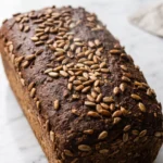 Schwarzbrot frisch gebacken — dunkles, kerniges Kastenbrot mit reichlich Sonnenblumenkernen auf weißem Marmoruntergrund.
