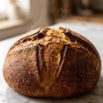Sauerteigbrot frisch gebacken — rundes, krustiges Landbrot mit kreuzförmigem Einschnitt auf weißem Marmor.