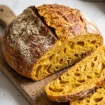 Saftiges Kürbisbrot mit Sauerteig frisch aus dem Gusseisentopf, angeschnitten auf rustikalem Holzbrett mit leuchtend oranger Krume.