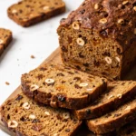 Früchtebrot mit Sauerteig und Vollkorn frisch gebacken in Kastenform, dick aufgeschnitten auf Holzbrett.