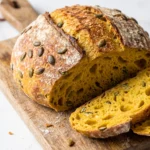 Kürbis-Dinkel-Sauerteigbrot frisch gebacken aus dem Gusseisentopf, angeschnitten auf Holzbrett mit leuchtend gelb-oranger Krume und Kürbiskernen.